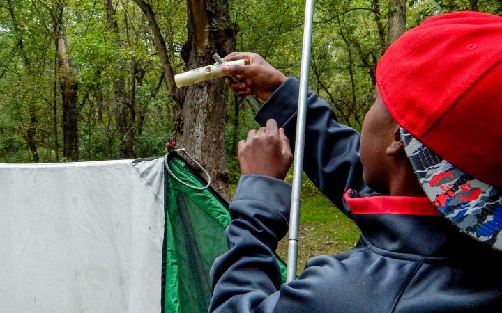 A young person works to tie a knot while assembling a tarp shelter in a wooded area. 